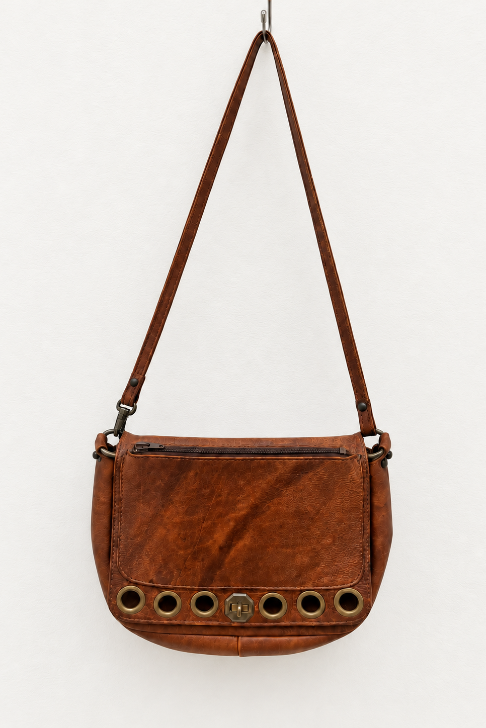 A brown leather shoulder bag with a long strap, featuring a front zip pocket and a row of circular metal grommets along the bottom edge, shown against a plain white background.