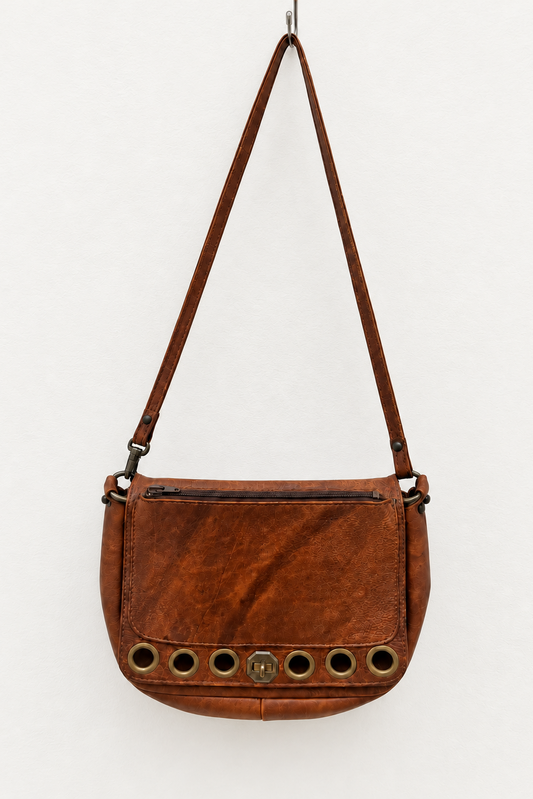 A brown leather shoulder bag with a long strap, featuring a front zip pocket and a row of circular metal grommets along the bottom edge, shown against a plain white background.