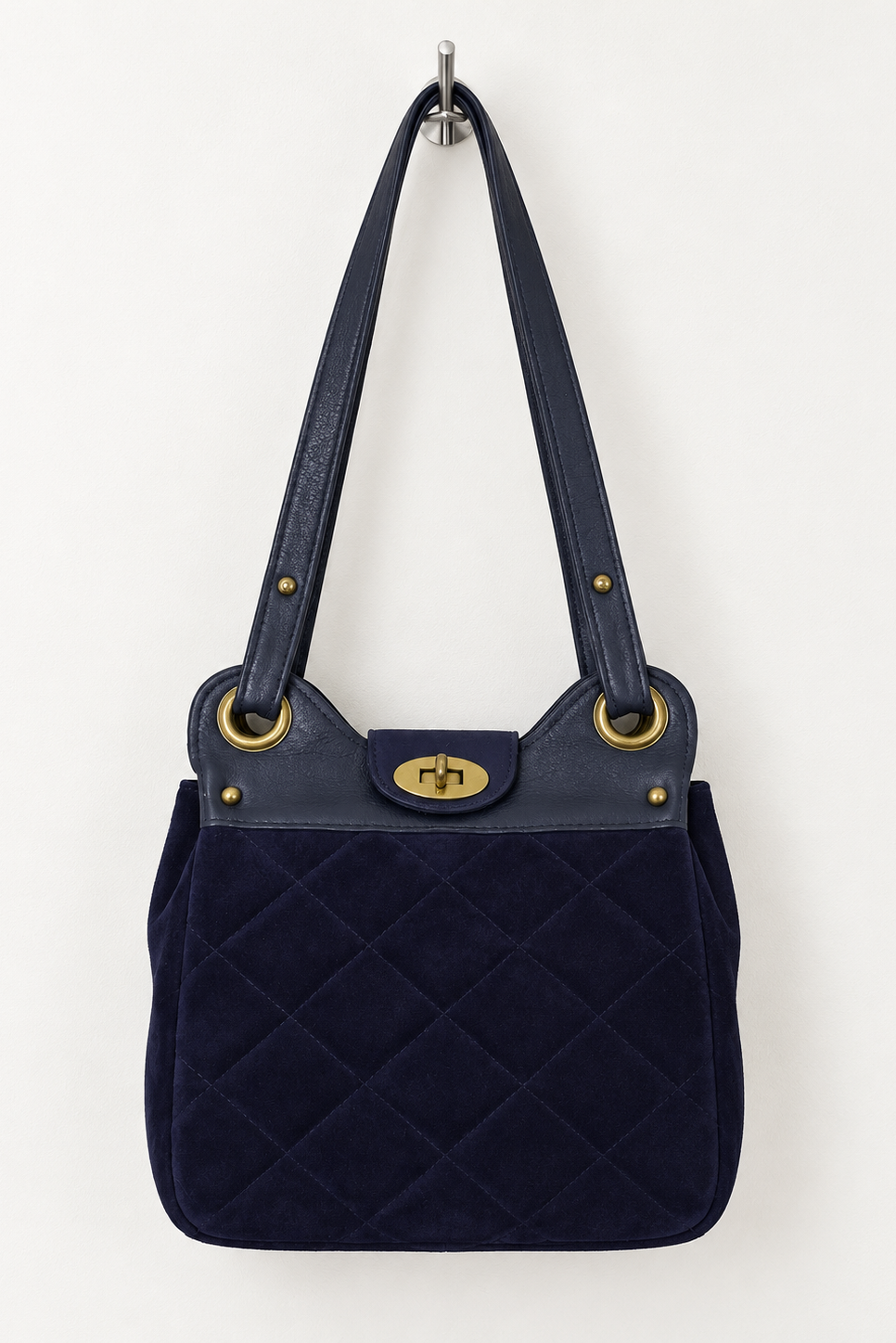 A navy blue quilted suede shoulder bag with dark blue leather straps and gold-tone hardware, hanging from a metal hook against a plain white wall.