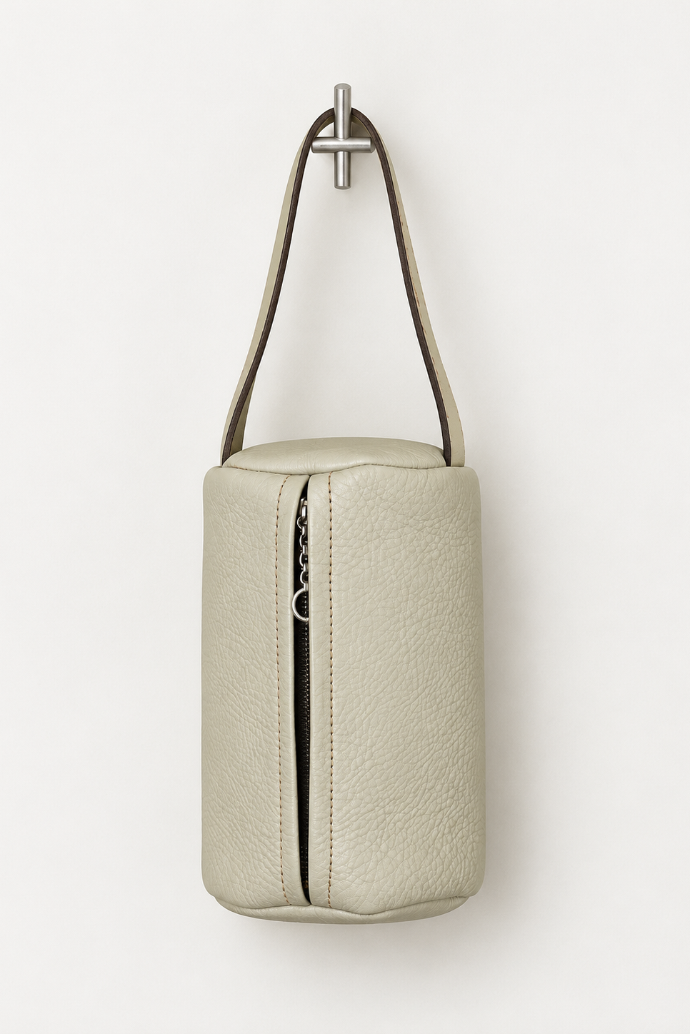 A light beige textured leather cylindrical pouch bag with a top zipper and a single shoulder strap, hanging from a silver wall hook against a plain white background.