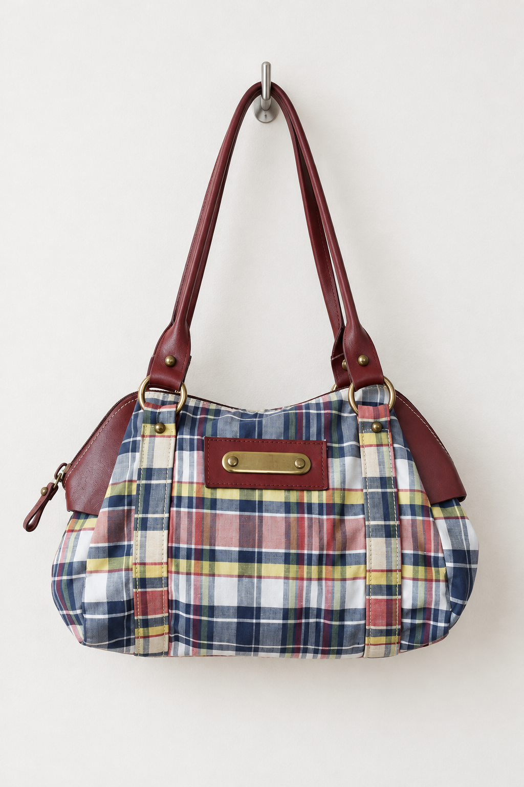 A plaid shoulder bag with reddish-brown leather handles and trim, hanging from a metal hook against a plain off-white wall. The bag features a blue, red, yellow, and white plaid pattern with a gold-tone metal clasp on the front.