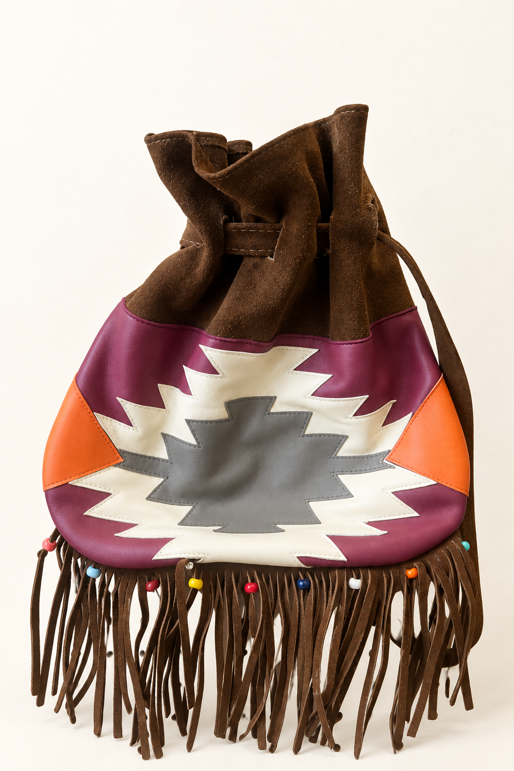 A drawstring pouch bag featuring a brown suede top and fringe, with a geometric patterned body in purple, orange, white, and gray, set against a plain off-white background.